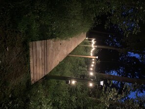 A wooden pathway leading through a wooded area with string lights along the path.
