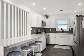 A modern kitchen with white cabinetry, a stainless steel refrigerator, a microwave, and a sink. There are three stools with patterned seats.