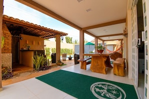 Une terrasse couverte avec une table et des chaises en bois, un paillasson vert et un mur en briques.