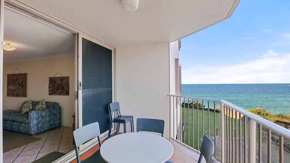 A balcony with a view of the sea, a dining table with chairs, and a sofa with cushions.