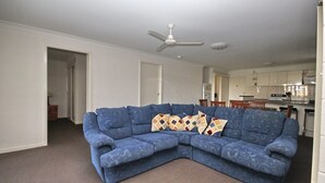 A living room with a blue sectional sofa, a ceiling fan, and a kitchen in the background. - Frasers Cottage - close to Laurieton shops and club (LAURIETON)