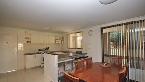 A compact kitchen with white cabinets, a microwave, and a stove. There is a dining table with chairs and a window with vertical blinds. - Frasers Cottage - close to Laurieton shops and club (LAURIETON)