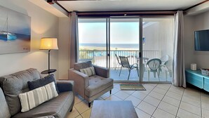 A hotel room with a sofa, a coffee table, a TV, and a balcony with a view of the beach.