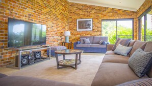 A living room with a brick wall, a flat-screen TV, a sofa, a coffee table, and a window with a view of greenery. - Aquila - 4 bedroom home with amazing valley views and swimming pool (Mount View)