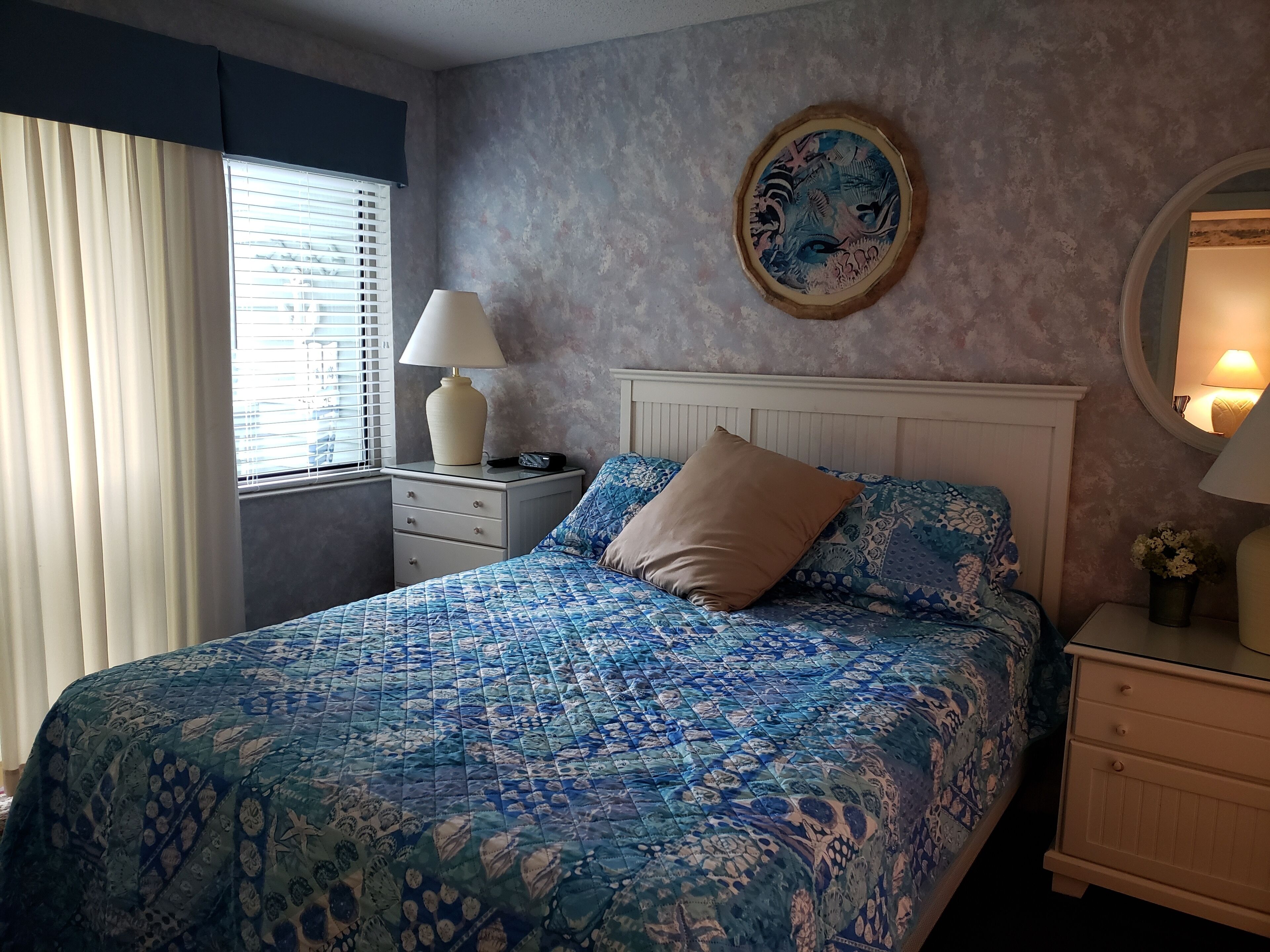 A bedroom with a bed, a bedside table, a lamp, a window with curtains, and a wall with a decorative plate.