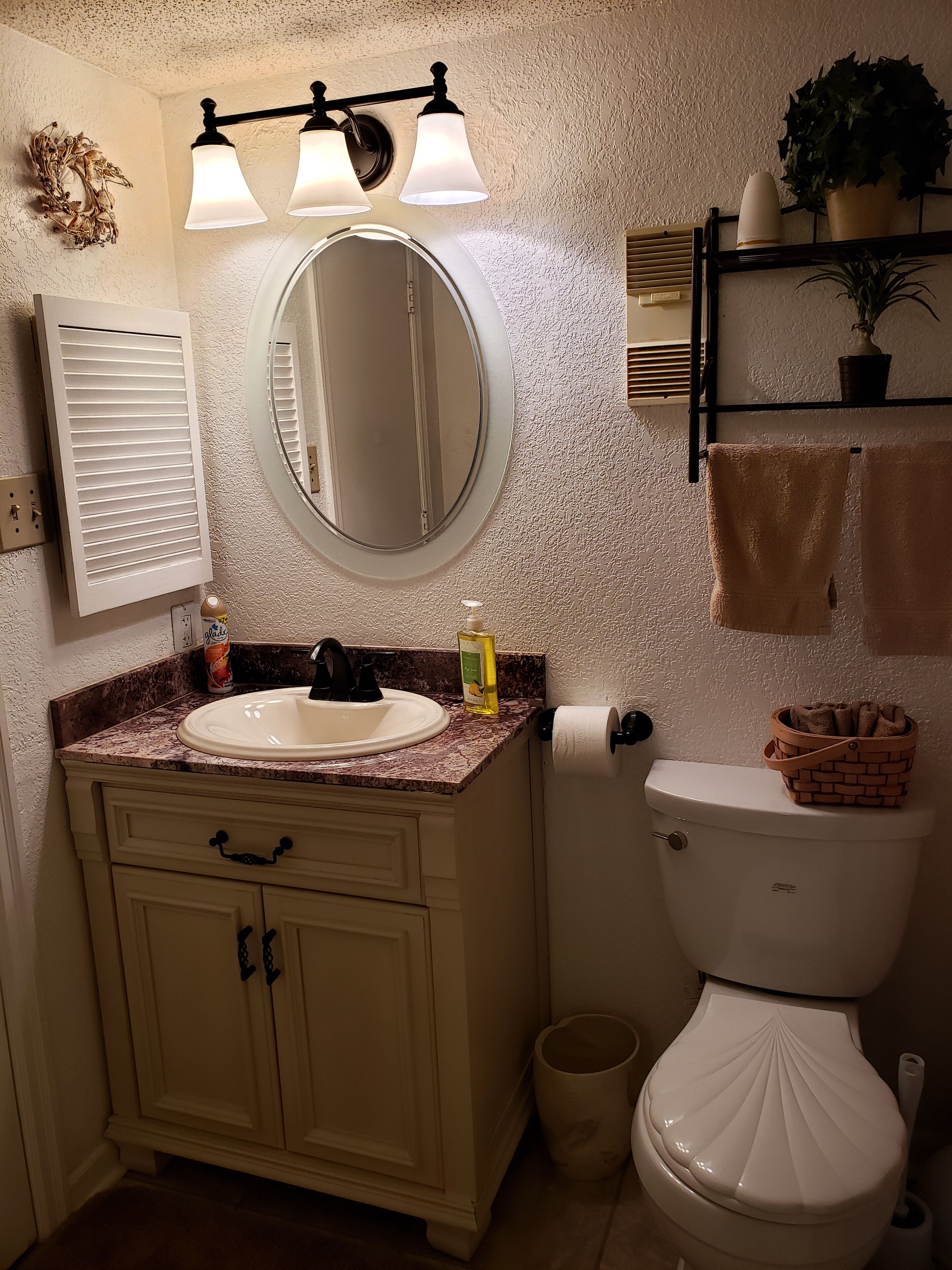 A bathroom with a white sink, a round mirror, a toilet, and a shelf with towels and a potted plant.