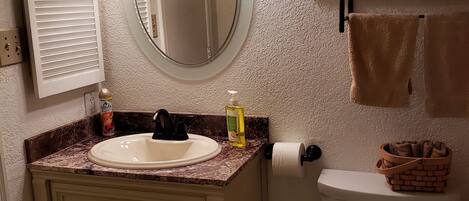 A bathroom with a white sink, a round mirror, a toilet, and a shelf with towels and a potted plant.
