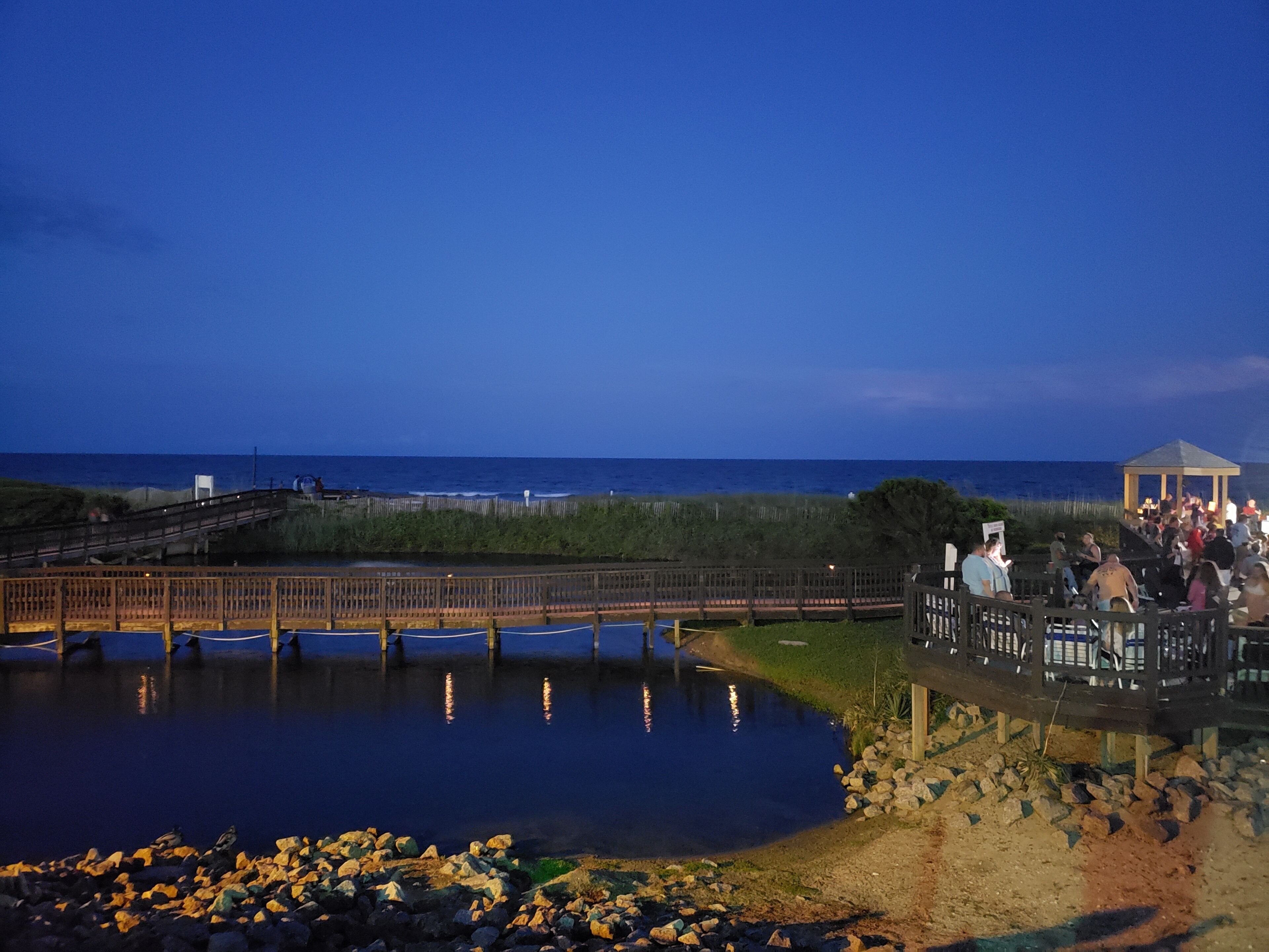 A coastal scene with a wooden bridge over a calm body of water, a gazebo, and a crowd of people.