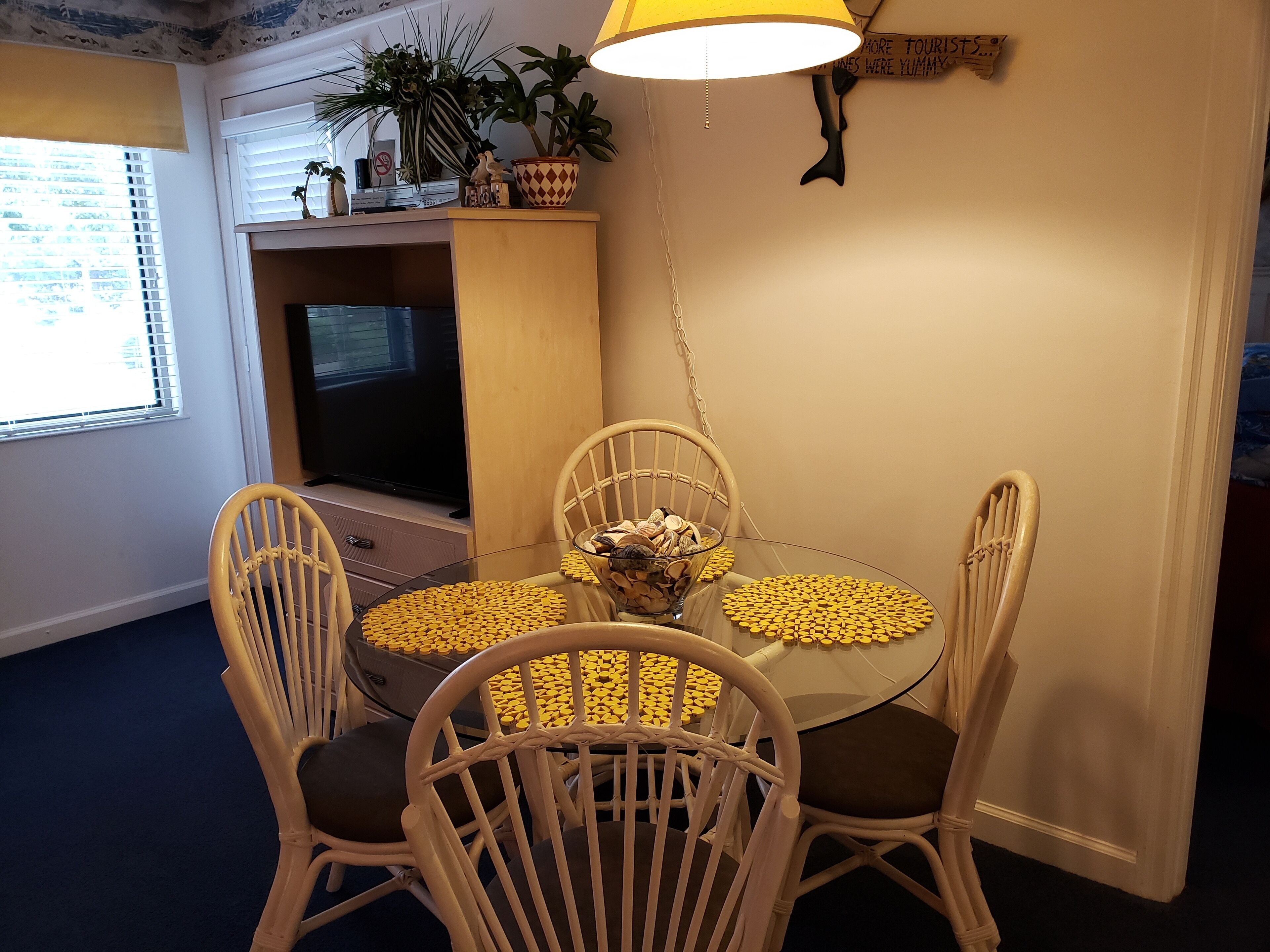 A dining area with a round glass table, four wicker chairs, yellow placemats, a potted plant, and a mounted animal head on the wall.