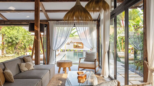 A modern living room with a glass coffee table, a sofa, and a chair. There is a pool visible through the glass doors.