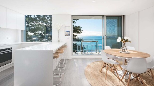 A modern kitchen with a large island, a dining area with a round table and chairs, and a view of the sea through large windows. - Cronulla Ocean View Apartment 87 (Cronulla)