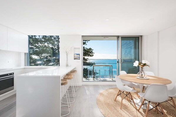 Una cocina moderna con una isla grande, un comedor con mesa redonda y sillas, y vistas al mar a través de amplios ventanales.