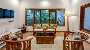 A living room with a sofa, two armchairs, a coffee table, and a floor lamp. There are large windows with a view of greenery outside.