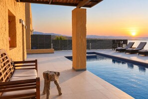 A poolside area with a view of the sunset, a wooden bench, and lounge chairs.