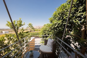 Un balcón con mobiliario de mimbre, una pequeña mesa y vistas a un jardín ajardinado con árboles y montañas lejanas.