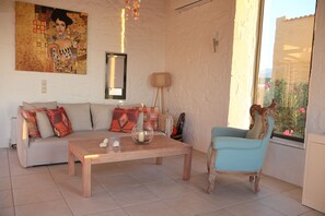 Una sala de estar con sofá, mesa de centro de madera, silla, un cuadro en la pared y ventana con vistas.