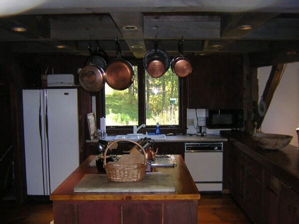 A kitchen with a central island, hanging pots, a refrigerator, a microwave, and a window with a view of greenery. - By Manchester, VT-Circa 1840 Hand Hewn Barn on private pond close to skiing. (Winhall)