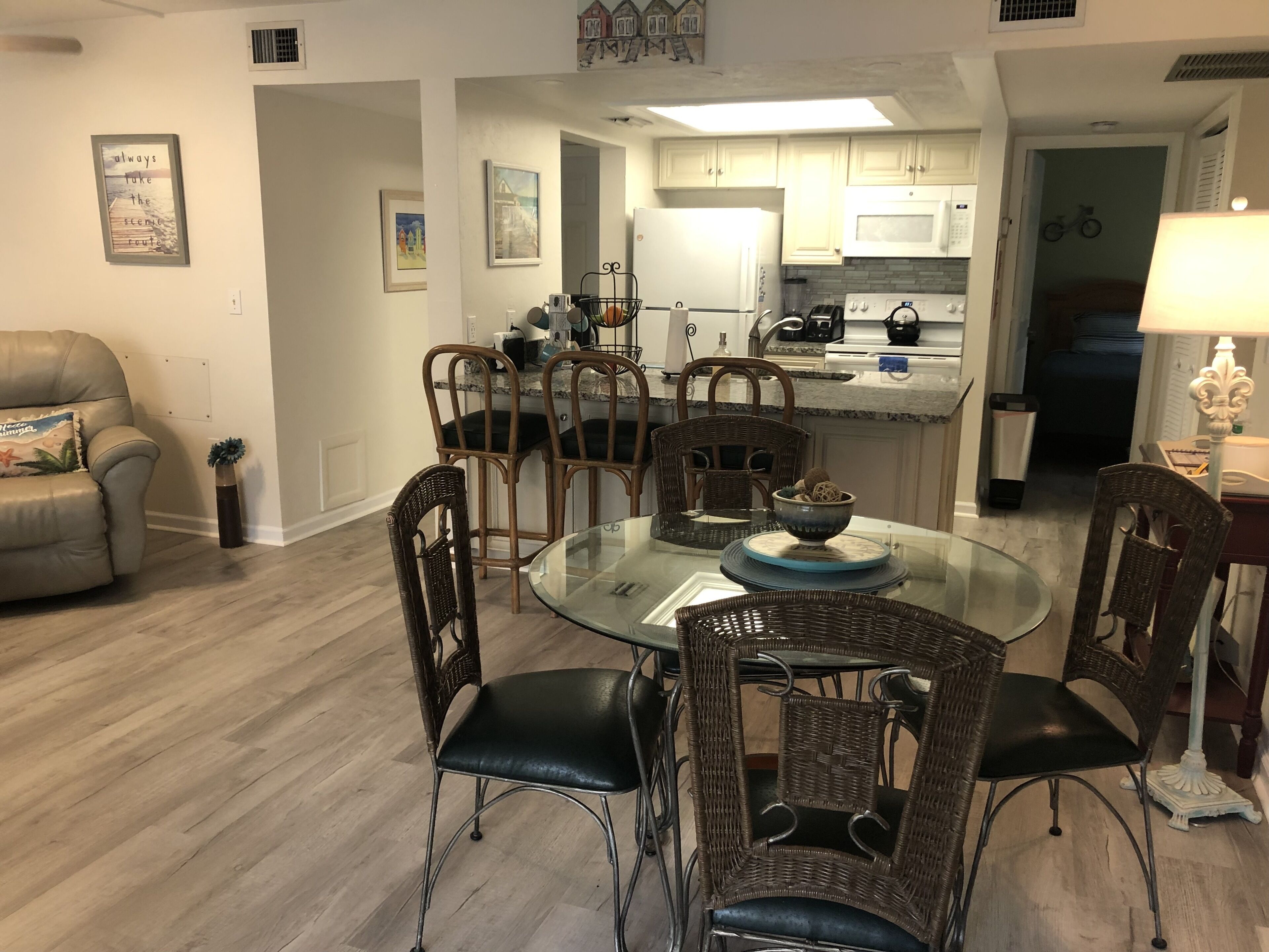 A dining area with a glass table surrounded by wicker chairs, a kitchen in the background, and a living room with a sofa and a lamp.