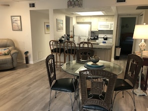 Ein Essbereich mit einem Glastisch, umgeben von Korbstühlen, eine Küche im Hintergrund und ein Wohnzimmer mit einer Couch und einer Lampe.