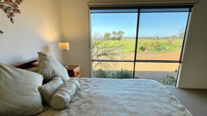 A bedroom with a large bed, a nightstand with a lamp, and a view of a green landscape through a window. - Al-Pac-Em Inn - Farm Stay (Kingscote)