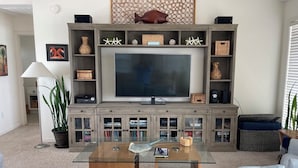 A living room with a large flat-screen TV, wooden shelving with decorative items, a glass coffee table, and a white sofa. - Terrapin Station: Oceanfront Home for Vacation Rental, Pet Friendly, Sleeps 10 (Surf City)