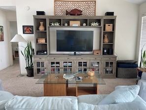 A living room with a large flat-screen TV, wooden shelving with decorative items, a glass coffee table, and a white sofa.