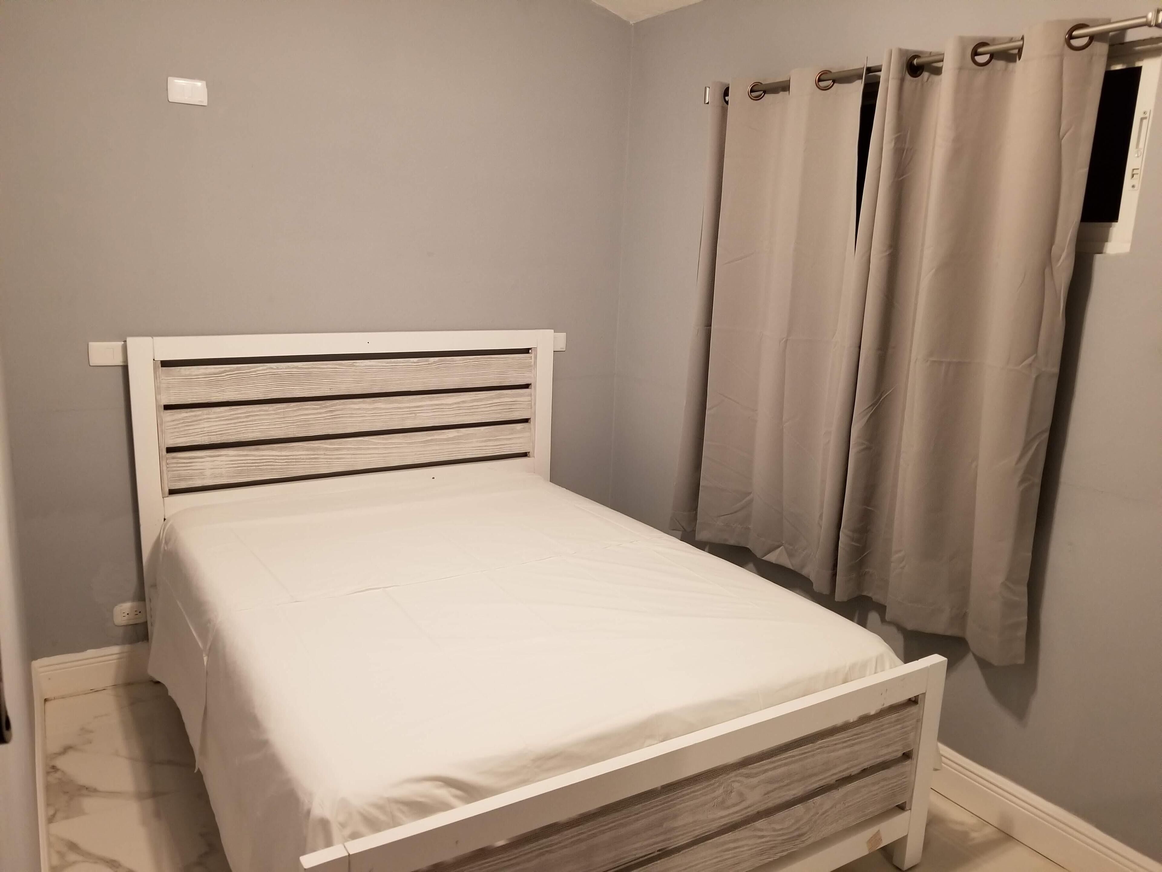 Una habitación pequeña con cama individual, cabecera de madera, colchón blanco sencillo y ventana con cortinas color beige.