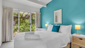 Habitación de hotel con una cama grande, dos mesitas de noche, un ventanal con cortinas, una pared de acento color turquesa y vistas a zonas verdes.