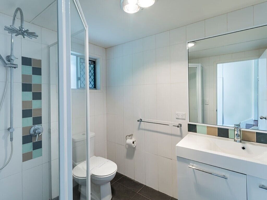 Et badeværelse med glasvæg omkring bruseren, toilet, vask med spejl og håndklædestang.