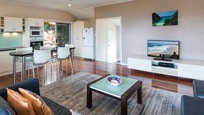 A modern living room with a TV, seating, and a kitchen area. - Family beach house across the road from Cylinder Beach. (Point Lookout)