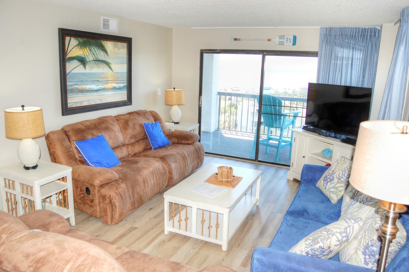A living room with a brown leather sofa, two blue cushions, a white coffee table, a television, and a balcony with blue chairs.