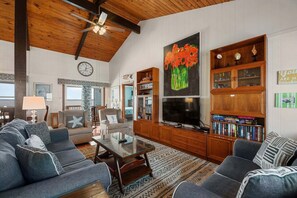 A spacious living room with a high ceiling, a large TV, and a bookshelf filled with books. - 419 Large Private Pier and Gorgeous Water Views (Nags Head)