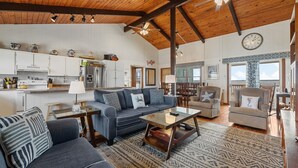 A spacious living area with a high ceiling, a kitchen with white cabinets, a dining area with a table and chairs, and a large window providing natural light.