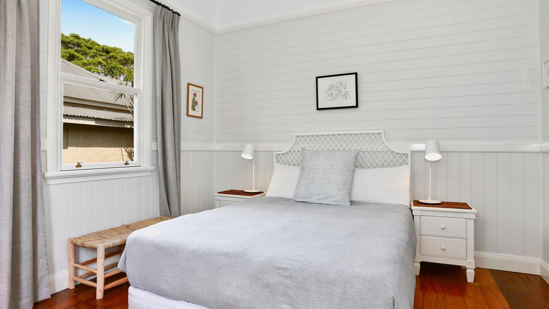 Un dormitorio con cama, mesitas de noche, ventana con cortinas, un cuadro enmarcado en la pared y un banco de madera.