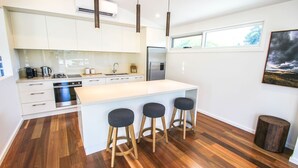 A modern kitchen with white cabinets, stainless steel appliances, a large island with stools, and a window with a view of trees. - VILLA URBANE : Bright (Bright)
