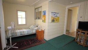 A small, single-room accommodation with a bed, a fan, a small table, a TV, and a bookshelf.