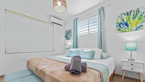 Un dormitorio con cama, una banca, mesita de noche, aire acondicionado de pared y ventana con cortinas.