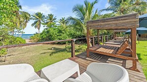 Una terraza de madera con sillas de mimbre blanco y una pérgola de madera con vistas a la playa.
