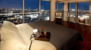 A bedroom with a large bed, a bedside table with a lamp, a tray with a glass of wine, and a view of a cityscape at night. - Amazing 2 BR/2.5 BA, 29/30th Floor Penthouse (Buenos Aires)