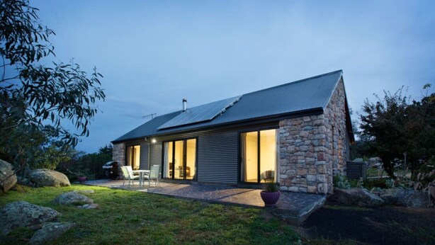 Uma casa moderna com fachada de pedra, grandes portas de vidro deslizantes e um pátio com mobiliário externo.