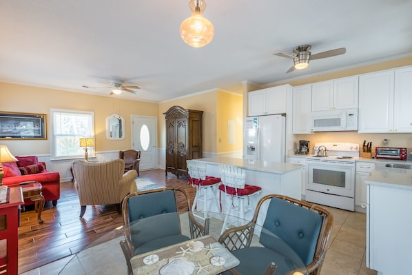A spacious living room with a red sofa, a glass coffee table, and a dining area with wicker chairs. There is a kitchen with white cabinets and a microwave.