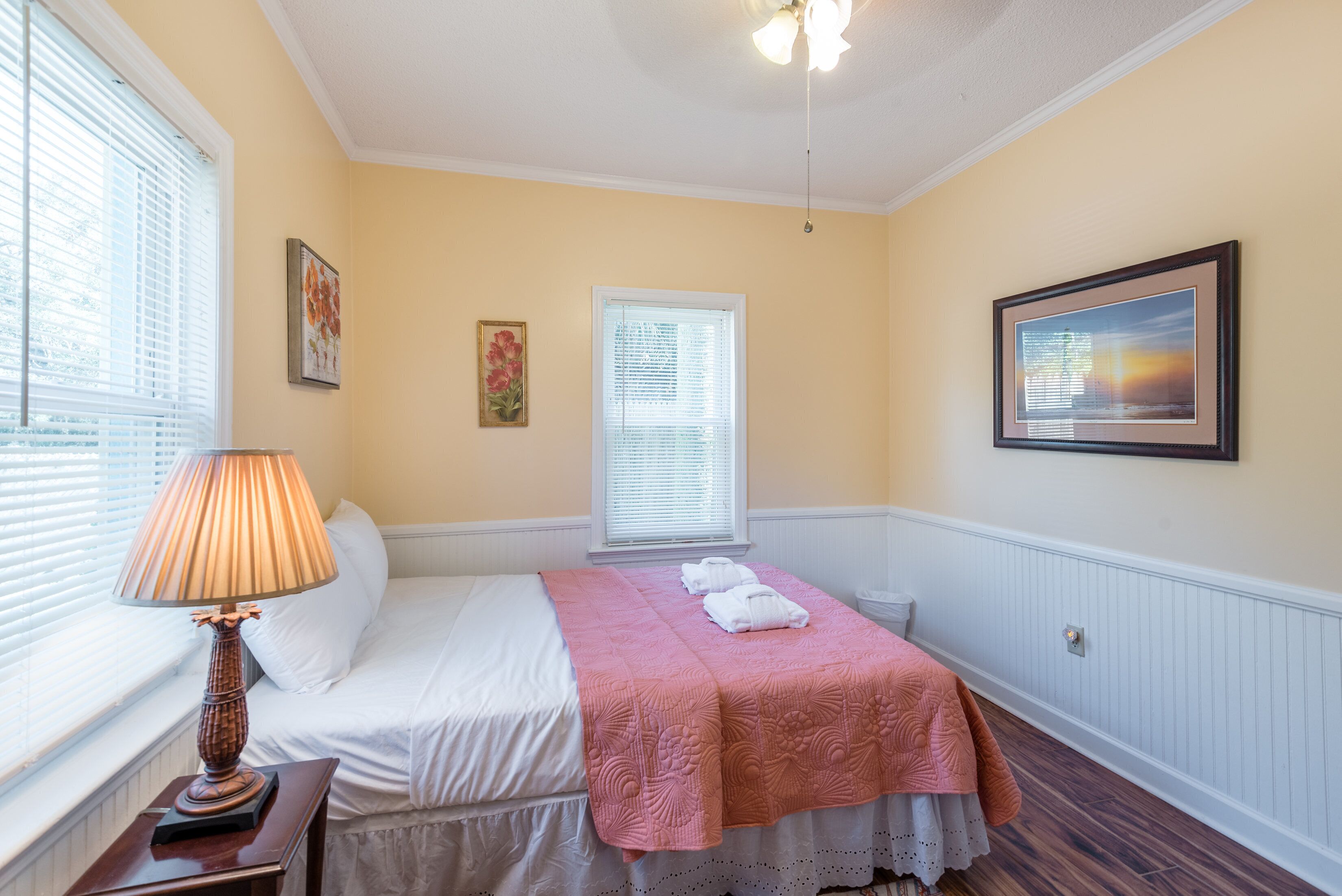 A hotel room with a bed, a bedside lamp, a window with blinds, a framed picture on the wall, and a ceiling fan.