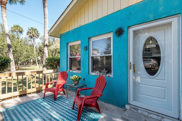 Une maison turquoise avec une porte blanche, deux chaises Adirondack rouges, une petite table et un pot de fleurs sur le porche.