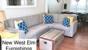 Ein Wohnzimmer mit einem grauen Ecksofa, gemusterten Kissen, einem Vintage-Koffer und einem Essbereich im Hintergrund.