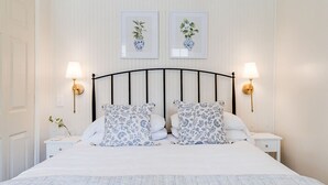 Een net opgemaakt bed met een wit- en blauw gebekeerd sprei, met aan weerszijden nachtkastjes met lampen, en botanische kunstwerken aan de muur.