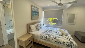 A bedroom with a bed, a nightstand, a ceiling fan, a window with a view of palm trees, and a painting on the wall. - Renovated Beach House in the Heart of Mooloolaba (Mooloolaba)