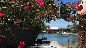 An outdoor patio with a pool, wooden furniture, and a view of a building across the water. - Renovated Beach House in the Heart of Mooloolaba (Mooloolaba)