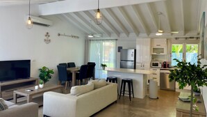A modern living room with a sofa, TV, and dining area. There is a kitchen with a fridge, microwave, and cabinets. The room has a high ceiling with exposed beams and pendant lights. - Renovated Beach House in the Heart of Mooloolaba (Mooloolaba)