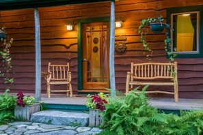 A wooden cabin with a porch, two wooden benches, a door with glass panels, and a hanging plant. - Unique Nature Inspired Home in Woods next to Lake (Mt Desert)