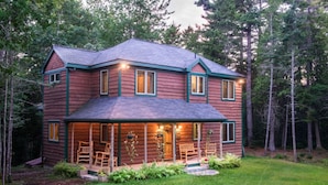 A two-story log cabin with a porch, multiple windows, and a well-maintained lawn. - Unique Nature Inspired Home in Woods next to Lake (Mt Desert)